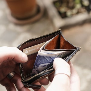 Op de afbeelding: Een bruine leren portemonnee met rits, met een zilveren kaartje erin en wat briefjes die eruit steken. De portemonnee is open en wordt vastgehouden door iemands handen.