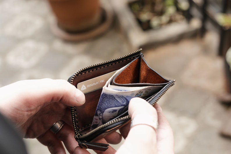 Op de afbeelding: Een bruine leren portemonnee met rits, met een zilveren kaartje erin en wat briefjes die eruit steken. De portemonnee is open en wordt vastgehouden door iemands handen.