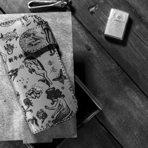 May include: A black and white leather phone case with a variety of tattoo-style designs, including a mermaid, a dolphin, a diamond, and a butterfly. The case has the text "新年快樂" in Chinese characters.