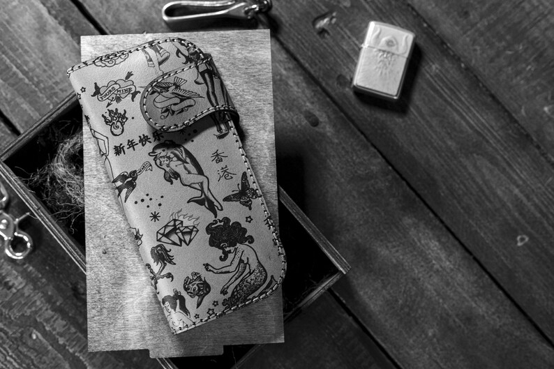 May include: A black and white leather phone case with a variety of tattoo-style designs, including a mermaid, a dolphin, a diamond, and a butterfly. The case has the text "新年快樂" in Chinese characters.