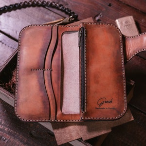 May include: A brown leather wallet with a zippered pocket and a chain attached. The wallet is open and the inside is visible. The wallet has the text "Grash Handmade in Georgia" on the inside.