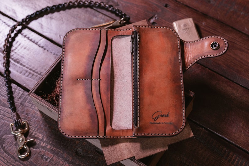 May include: A brown leather wallet with a zippered pocket and a chain attached. The wallet is open and the inside is visible. The wallet has the text "Grash Handmade in Georgia" on the inside.