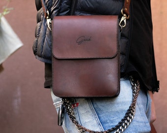 Può includere: Una borsa a tracolla in pelle marrone con una catena argentata attaccata alla parte inferiore. La borsa ha una chiusura a lembo e una piccola tasca sul davanti. La borsa ha la parola "Grash" impressa sul davanti.
