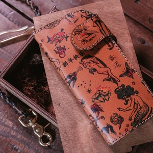 May include: A brown leather wallet with a colorful tattoo-style design featuring mermaids, dolphins, and other nautical imagery. The wallet has a flap closure and a chain attached to it.