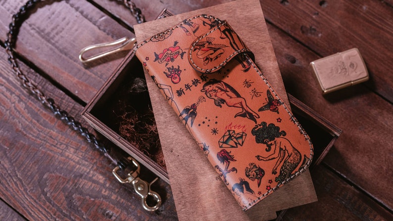 May include: A brown leather wallet with a colorful tattoo-style design featuring mermaids, dolphins, and other nautical imagery. The wallet has a flap closure and a chain attached to it.