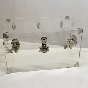 May include: A clear acrylic storage trunk with silver metal clasps and hinges. The trunk is empty and has a rectangular shape.