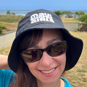 May include: A woman wearing a black bucket hat with the text "MAUI GIRL" in white, and sunglasses. She is wearing a turquoise t-shirt with the text "MAUI SURFER GIRLS" in white. The background shows a sunny outdoor scene.
