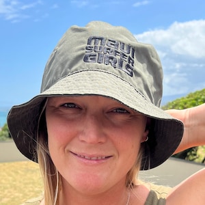 May include: An olive green bucket hat with the text "MAUI SURFER GIRLS" in black is worn by a person. The person is also wearing an olive green tank top and a silver necklace with a small pendant. The background includes a blue sky and green foliage.