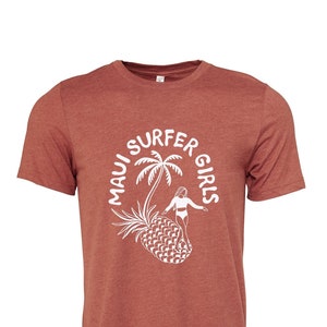 May include: A rust-colored t-shirt with a white graphic of a woman surfing on a pineapple with a palm tree behind her. The graphic is in a circle with the text "Maui Surfer Girls".