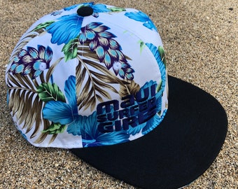 Può includere: Cappellino da baseball bianco e blu con motivo floreale e visiera nera. La parte anteriore del cappello ha il testo "Maui Surfer Girls" in blu.