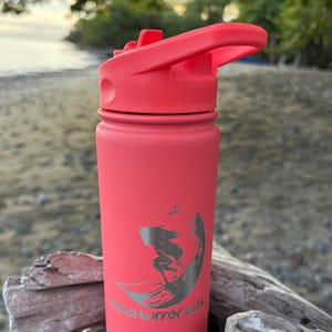 Puede incluir: Una botella de agua de color coral con tapa abatible. La botella presenta un gráfico plateado de un surfista y el texto "Maui Surfer Girls". La botella es cilíndrica y parece ser de metal.