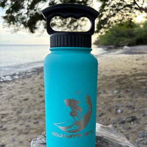 Puede incluir: Una botella de agua turquesa con asa y tapa negras. La botella presenta un gráfico plateado de un surfista sobre una ola, con las palabras "maui surfer girls" debajo. La botella está sobre madera flotante.