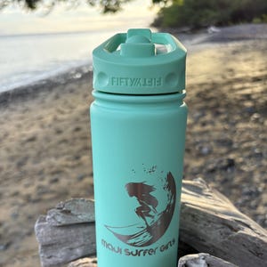 Puede incluir: Una botella de agua turquesa con tapa abatible. La botella tiene el texto "FIFTY/FIFTY" en la tapa y un gráfico de una surfista con el texto "maui surfer girls". La botella está sobre madera flotante.