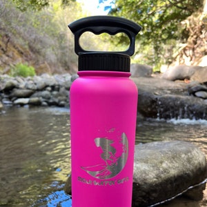 Puede incluir: Una botella de agua de acero inoxidable de color rosa brillante con asa y tapa negras. La botella tiene un logotipo plateado que dice "Maui Surfer Girl" con una silueta de una surfista.