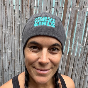 Puede incluir: Un gorro gris con el texto "Maui Surfer Girls" bordado en azul turquesa.