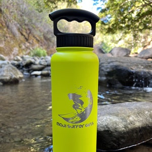 Puede incluir: Una botella de agua de color amarillo brillante con un asa negra y un logotipo plateado que dice "Maui Surfer Girls" con una silueta de una surfista en una ola.