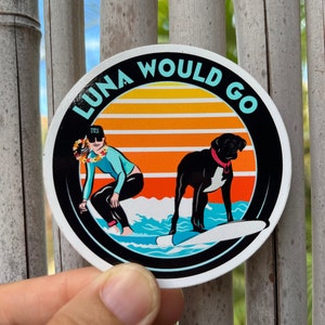 May include: A round sticker with a black border and a white background. The sticker features a sunset with orange, yellow, and red stripes. A woman in a blue shirt and black pants is surfing on a surfboard with a black dog. The text "LUNA WOULD GO" is written in a black circle around the image.