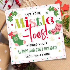 May include: A Christmas gift tag with the text "For Your Mistle Toes!" and festive illustrations of socks and holly. The tag is placed on a wrapped gift with a red snowflake decoration and sprigs of evergreen.
