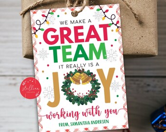 以下が含まれることがあります： 赤い文字と緑の文字で「We make a great team. It really is a joy working with you. From, Samantha Anderson」と書かれた白いクリスマスギフトタグ。タグは、雪片、ライト、ベルのついたリースなど、お祝いのデザインが施されています。