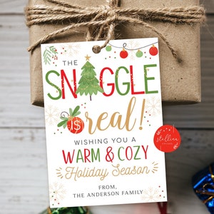 May include: A wrapped gift with a festive tag that reads "The Snuggle is Real!" with a Christmas tree illustration. The tag wishes a warm and cozy holiday season. The gift is tied with twine.