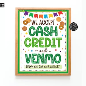 May include: Green and white sign with the text "We accept cash, credit and Venmo. Thank you for your support!" with a border of brown wood and colorful flags and cookie images.
