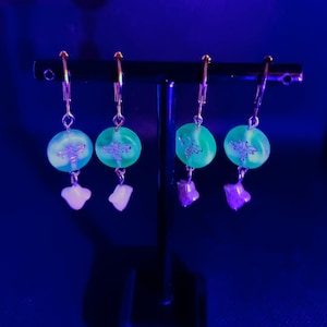 May include: Four pairs of green and white dangle earrings with a small crystal charm hanging from each earring. The earrings are displayed on a black stand.
