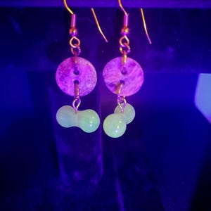 May include: A pair of handmade earrings. Each earring features a circular, textured, purple-toned stone, connected to a gold-colored hook. Below the stone hang two light green, bead-like shapes. The earrings are displayed against a dark background.