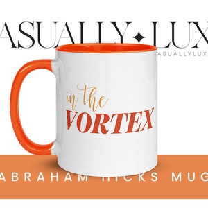 May include: White ceramic mug with an orange rim and handle. The mug has the text "in the vortex" printed in red and gold on the front. The mug is sitting on a light orange surface with the text "Abraham Hicks Mug" printed below the mug.