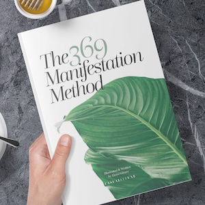 以下が含まれることがあります： 黒い文字で「The 369 Manifestation Method」というタイトルが書かれた白い本。本は緑の葉で飾られており、下部に「Illustrated & Written by Elena Gliturr CASUALLY LUXE」という文字が書かれている。本は手で持たれている。