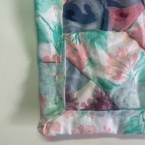May include: Close-up of a quilted fabric with a floral pattern. The fabric features a patchwork design with various floral prints in shades of pink, blue, and green. The edges are neatly sewn, showcasing the craftsmanship of the quilt.