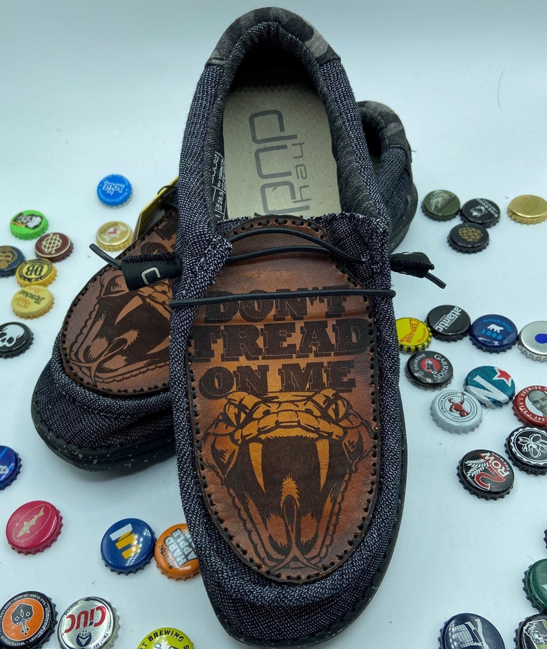 May include: A pair of gray canvas slip-on shoes with a brown leather patch featuring a snake and the text "Don't Tread On Me".