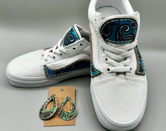 Puede incluir: Zapatillas de lona blancas con detalles paisley azules y ribetes de cuero marrón. Pendientes a juego en forma de lágrima con diseño paisley verde y negro se muestran junto a los zapatos. Las zapatillas tienen cordones y suelas blancas.