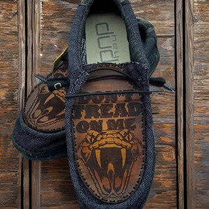 May include: A pair of black canvas shoes with brown leather accents. The shoes feature a snake graphic and the text "Don't Tread On Me".