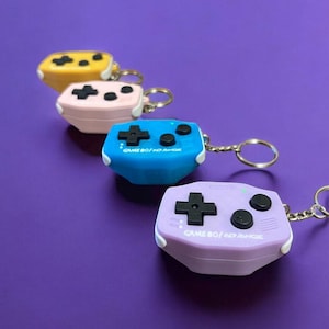 Op de afbeelding: Vier Game Boy-sleutelhangers in verschillende kleuren. De sleutelhangers zijn geel, roze, blauw en paars. Elke sleutelhanger heeft de tekst "GAME BOY/NO RANGE" erop gedrukt.