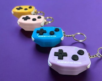 May include: Four different colored Game Boy keychain replicas. The keychains are in yellow, pink, blue, and purple. Each keychain has the text "GAME BOY/NO RANGE" printed on it.