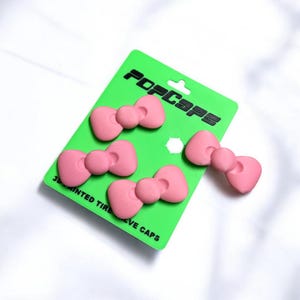 Peut inclure: Quatre bouchons de valve de pneu roses en forme de nœud papillon sur une carte vert vif. La carte porte le texte "POPCAPS" en noir et "3D PRINTED TIRE VALVE CAPS" en dessous. Le fond est blanc.