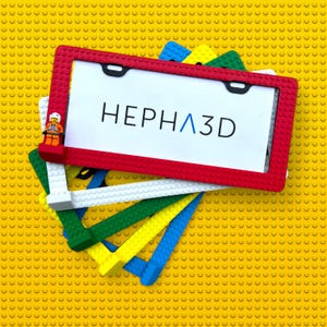 May include: A red license plate frame with the text "HEPHA3D" in black and blue. The frame is made of red plastic and is surrounded by other colourful frames. A small Lego figure is on the top left corner.