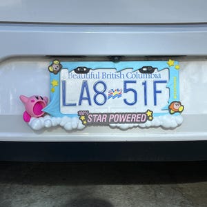 May include: A license plate frame with a light blue border and cartoon characters. The license plate reads "LA8 51F" and is from British Columbia. The frame has the text "100% STAR POWERED".
