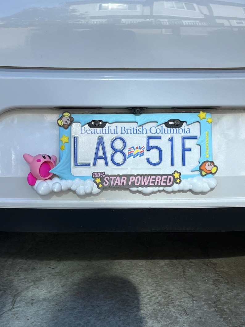 May include: A license plate frame with a light blue border and cartoon characters. The license plate reads "LA8 51F" and is from British Columbia. The frame has the text "100% STAR POWERED".