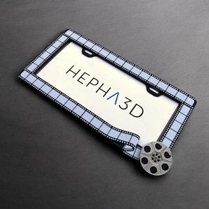 Könnte beinhalten: Ein schwarzer und weißer Filmstreifen-Kennzeichenrahmen mit dem Text "HEPHA3D" auf weißem Hintergrund. Eine silberne Filmrolle befindet sich in der unteren rechten Ecke des Bildes.