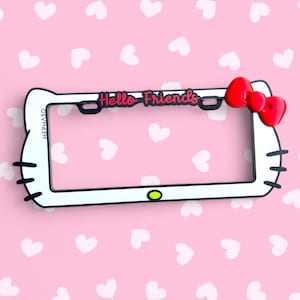 Peut inclure: Cadre de plaque d'immatriculation blanc avec un design Hello Kitty. Il présente un nœud rouge, des moustaches noires et les mots "Hello Friends" en rose. Fond rose avec des cœurs blancs.