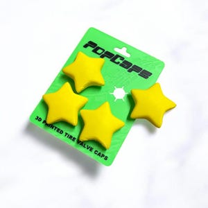 Puede incluir: Un paquete de cuatro tapones de válvula de neumático amarillos en forma de estrella. En una tarjeta verde con el texto "POP CAPS" y "3D PRINTED TIRE VALVE CAPS". Los tapones son de color amarillo brillante.