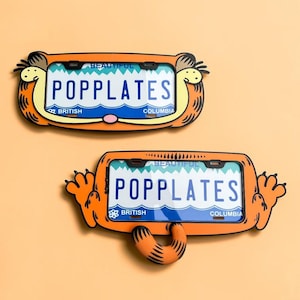 Op de afbeelding: Twee oranje kentekenplaathouders met een cartoon kat ontwerp. Beide houders tonen de tekst BEAUTIFUL, POPPLATES en BRITISH COLUMBIA. De bovenste houder toont een kattengezicht, de onderste toont een kat met uitgestrekte poten en een staart.