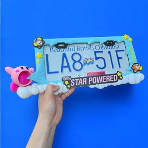May include: A decorative license plate frame with the text "Beautiful British Columbia" and the license plate number "LA8-51F". The frame is light blue with cartoon character accents and the words "STAR POWERED". A pink cartoon character is on the left.