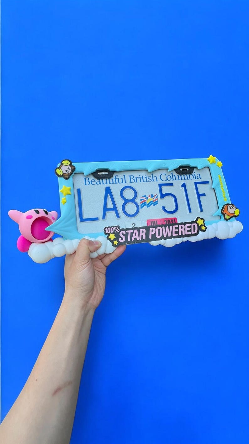 May include: A decorative license plate frame with the text "Beautiful British Columbia" and the license plate number "LA8-51F". The frame is light blue with cartoon character accents and the words "STAR POWERED". A pink cartoon character is on the left.