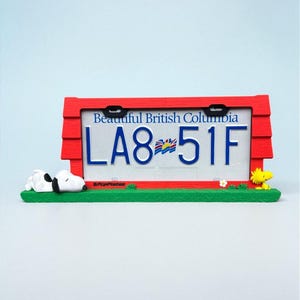 May include: A red license plate frame with the text "Beautiful British Columbia" and the license plate number LA8-51F. The frame has a green base with Snoopy and Woodstock figurines.