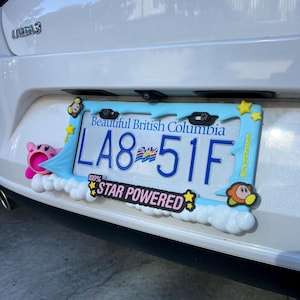 May include: A car license plate frame with a blue border and cartoon character accents. The license plate reads "LA8-51F" and has the text "Beautiful British Columbia" at the top. The frame also has the text "100% STAR POWERED".