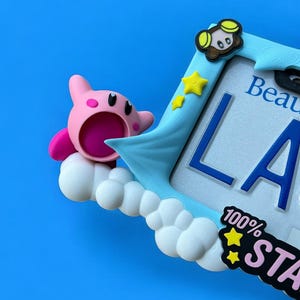 May include: A light blue picture frame with a cartoon character and the text "LA". The frame has a cloud and star design, with the words "100% STA" on the bottom right. The background is a solid blue.
