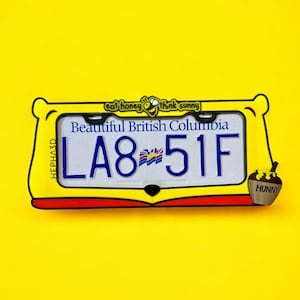 Può includere: Una cornice gialla per targa con la scritta "eat honey think sunny" e una targa che recita "Beautiful British Columbia" con il numero "LA8 51F". Un piccolo vasetto di miele con la scritta "Hunny" è a destra.