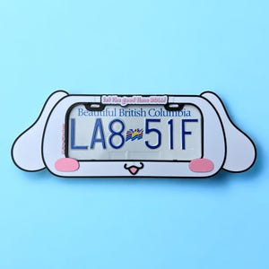 May include: A novelty license plate frame in the shape of a cartoon dog. The frame surrounds a license plate with the text "Beautiful British Columbia" and the number "LA8-51F". The background is light blue.